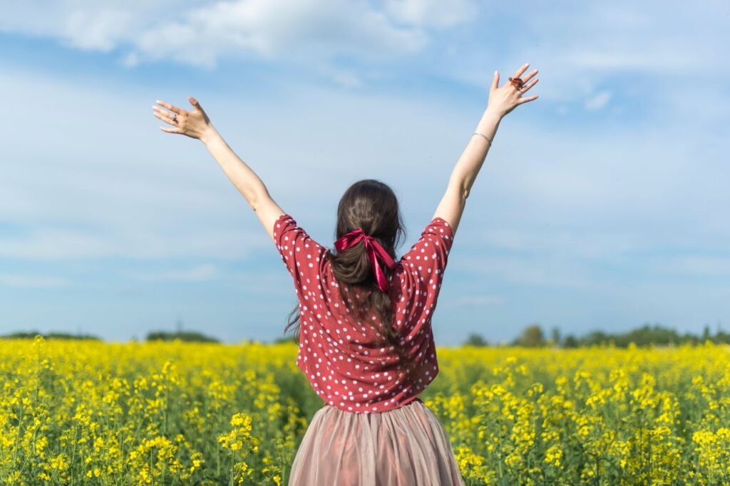 A woman in a polka dot shirt raising her arms in a bright yellow flower field, expressing freedom and joy.